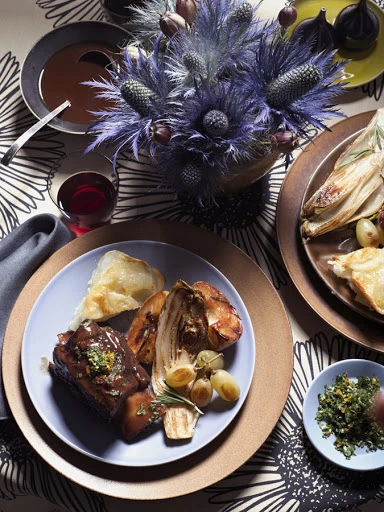 Overhead shot of a beautifully plated dish with braised short ribs, roasted vegetables, and herbs, set against a floral tablecloth.