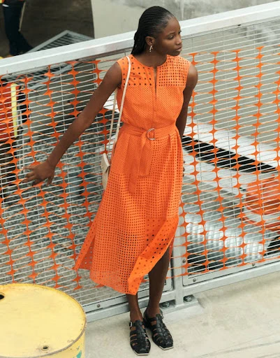 A slender Black woman poses near a metal railing, wearing a sleeveless orange dress with geometric cutouts, in a medium outdoor shot.