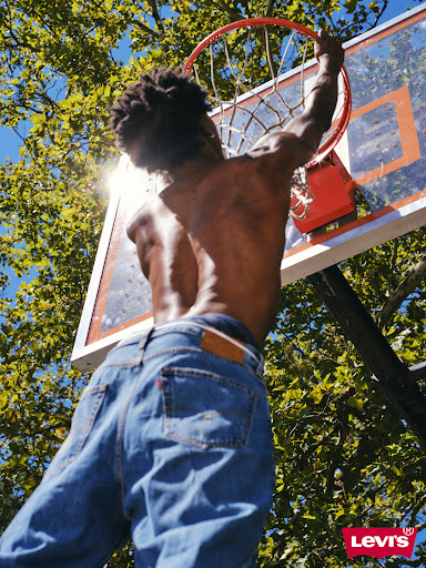A muscular adult male with dreadlocks hangs from a basketball hoop, wearing jeans in a sunny outdoor setting with leafy trees.