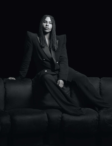 Portrait of a person with long braids wearing a double-breasted blazer and slacks sitting on a dark velvet couch against a black backdrop.
