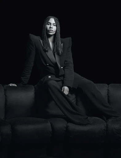 Portrait of a person with long braids wearing a double-breasted blazer and slacks sitting on a dark velvet couch against a black backdrop.