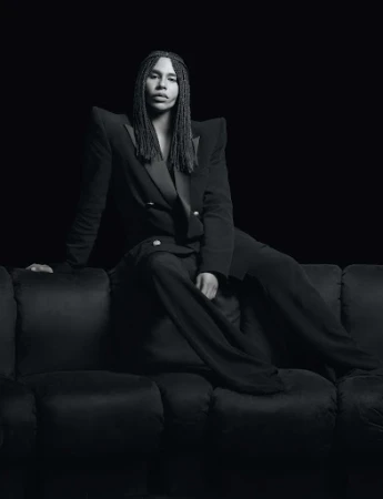 Portrait of a person with long braids wearing a double-breasted blazer and slacks sitting on a dark velvet couch against a black backdrop.