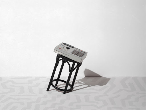 A vintage drum machine sits atop a black stool against a bright white backdrop with minimalist modern geometric patterns on the floor.
