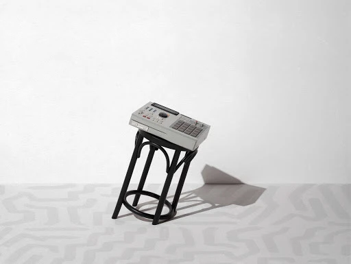 A vintage drum machine sits atop a black stool against a bright white backdrop with minimalist modern geometric patterns on the floor.