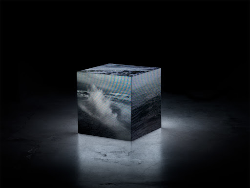 Abstract cube with ocean wave imagery on a dark marble surface, illuminated in a black studio setting, creating a surreal atmosphere.