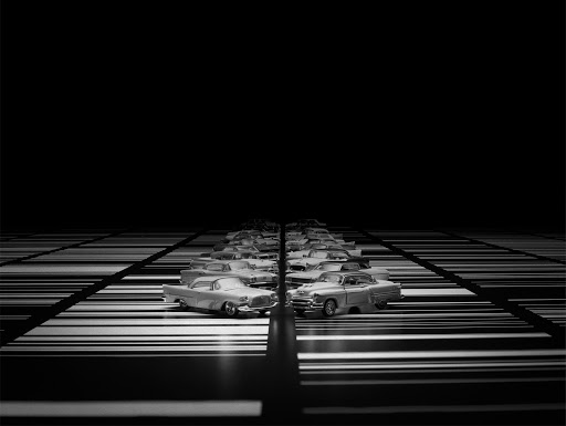 A long row of vintage automobiles displayed on a black and white striped surface receding into the dark background with low-key lighting.