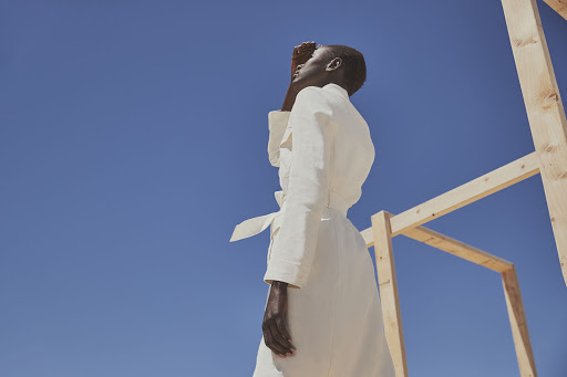 Adult female model in a long white coat poses with her hand shielding her face against a clear blue sky near a wooden frame structure.