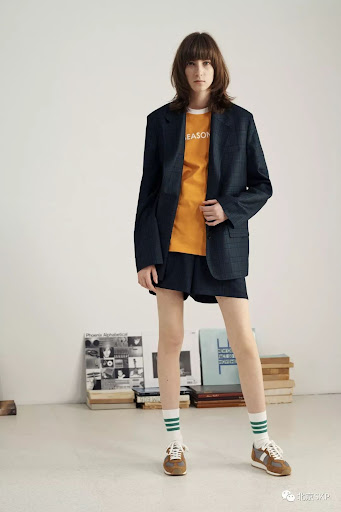 Adult female fashion model poses in an oversized blazer, shorts, socks, and sneakers in a studio setting with books nearby.