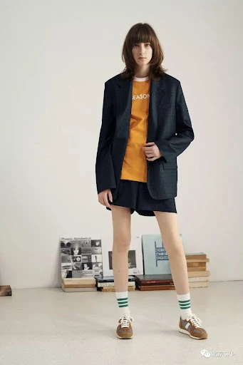 Adult female fashion model poses in an oversized blazer, shorts, socks, and sneakers in a studio setting with books nearby.