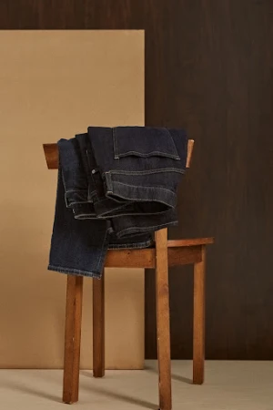 Folded dark denim jeans draped over a wooden chair against a split beige and brown panel background.