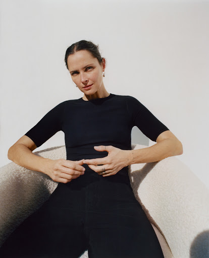 Portrait of an adult woman with dark hair, wearing black top and pants, seated in a white chair against a light background in a studio.