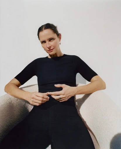 Portrait of an adult woman with dark hair, wearing black top and pants, seated in a white chair against a light background in a studio.