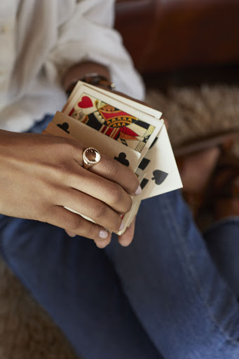 Person holding playing cards, wearing blue jeans and a white shirt indoors. A ring is visible on their left hand.