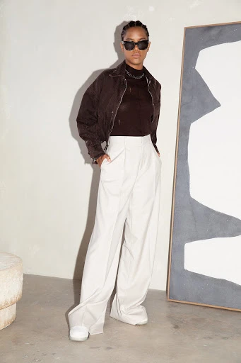 Woman in brown jacket, black shirt, and white pants poses near abstract art and stone stool, indoor studio lighting, fashion shoot.