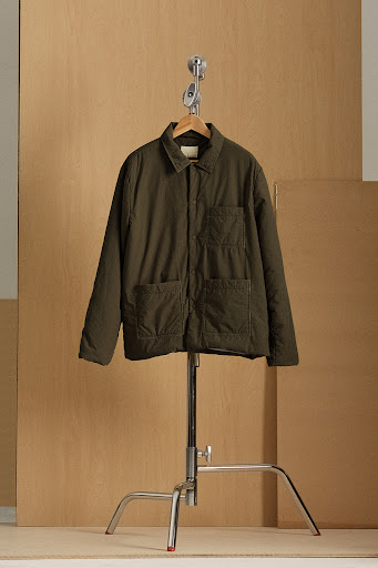 Olive green quilted jacket on a wooden hanger, suspended from a chrome stand against a light beige background.