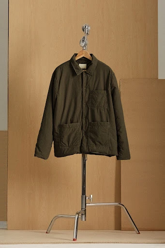 Olive green quilted jacket on a wooden hanger, suspended from a chrome stand against a light beige background.