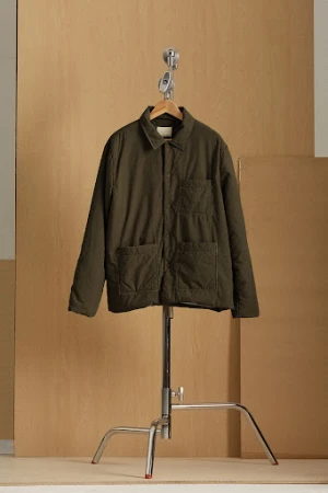 Olive green quilted jacket on a wooden hanger, suspended from a chrome stand against a light beige background.