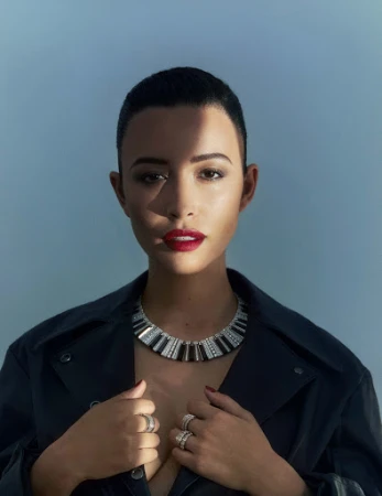 Close-up portrait of a woman wearing a statement diamond necklace and dark jacket against a pale background.
