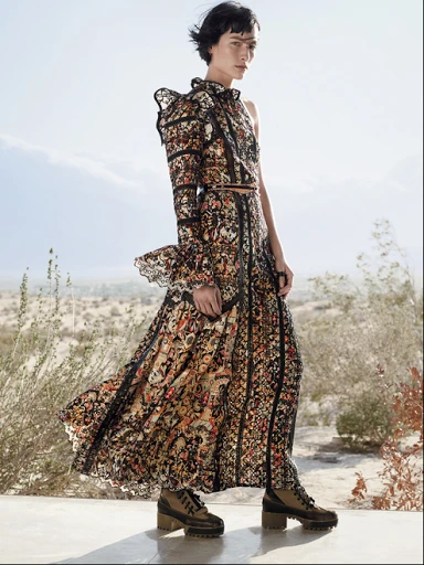 Fashionable woman in a floral, black, and tan dress and boots standing in a desert landscape with mountains in the distance.