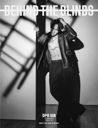 Black and white studio portrait of a shirtless adult male wearing a leather jacket and pants, posing against blinds and dramatic lighting.