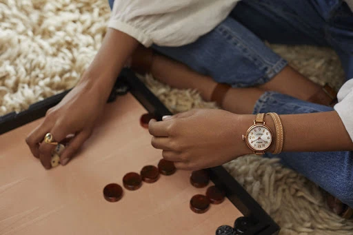 Woman wearing a wristwatch playing a board game indoors on a shag rug with game pieces, dice, and blue jeans in soft light.