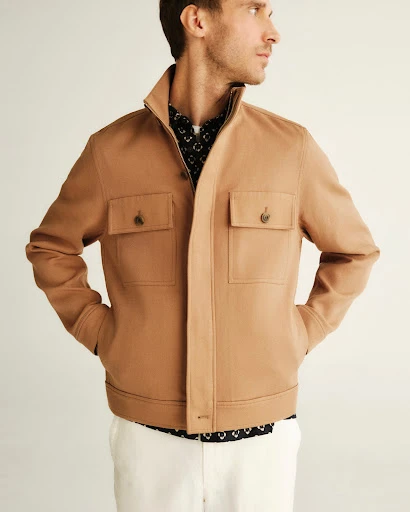 A fair-skinned man in a tan collared jacket and white pants poses against an off-white background, his gaze directed off-camera.