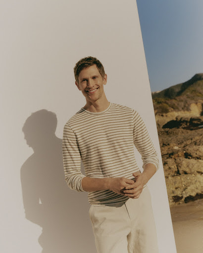 A man with short brown hair smiles wearing a striped shirt and tan pants, standing against a wall with a desert mountain backdrop.