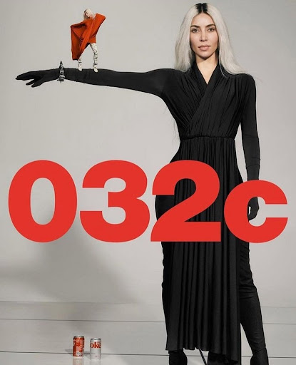 Adult woman in long black dress poses indoors with a small doll wearing an orange cape and two cans of Coca-Cola in the background.