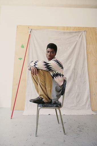 Young man wearing a patterned sweater and tan pants sits on a metal chair in a light-filled studio with neutral plywood backdrop.