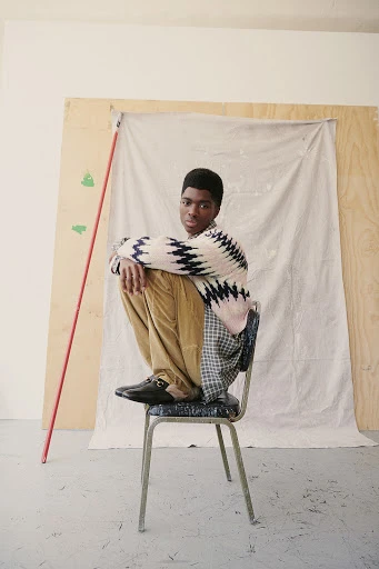 Young man wearing a patterned sweater and tan pants sits on a metal chair in a light-filled studio with neutral plywood backdrop.