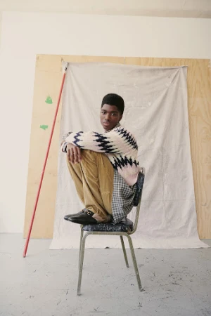 Young man wearing a patterned sweater and tan pants sits on a metal chair in a light-filled studio with neutral plywood backdrop.