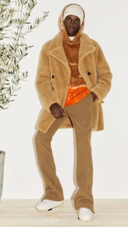Black man wearing a tan fuzzy double-breasted coat and matching trousers over an orange tie-dye hoodie.