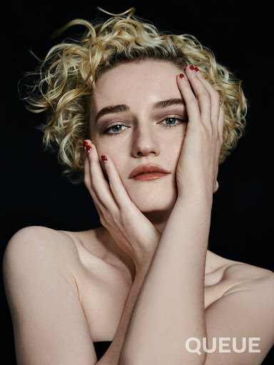 Close-up portrait of a young woman with blonde curly hair, red manicure, and light makeup, posing with her hands near her face.