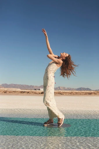 Woman in a dress stands on a shallow pool with arms raised against a clear blue sky and desert landscape in an outdoor fashion shot.