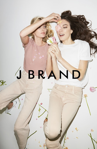 Two women pose with flowers in a light, airy studio setting, wearing casual tops and jeans. J Brand logo is visible in the center.