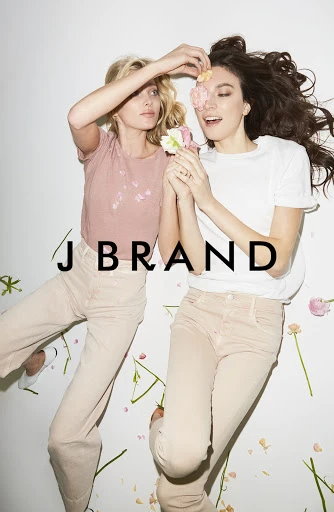 Two women pose with flowers in a light, airy studio setting, wearing casual tops and jeans. J Brand logo is visible in the center.