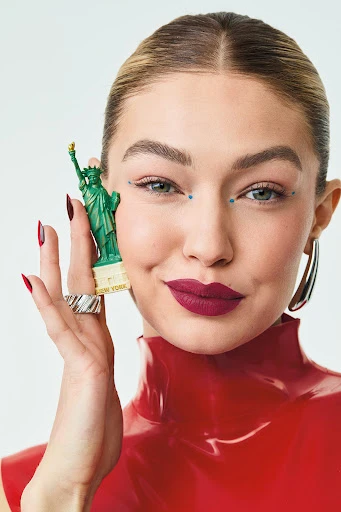 Portrait of an adult woman holding a miniature Statue of Liberty, wearing a red top, silver hoop earrings, and ring with a white background.