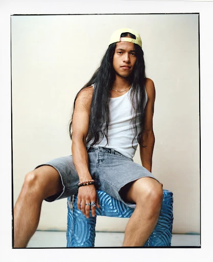 Young man with long hair poses indoors sitting on an ornate stool wearing a white tank top, gray shorts, and a yellow cap.