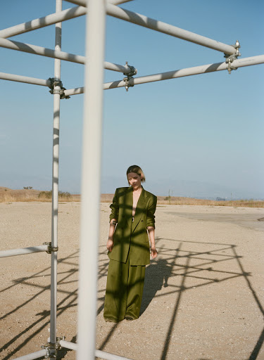 A woman poses in a green suit outdoors in a tan, arid landscape, framed by a white scaffolding structure against a clear blue sky.