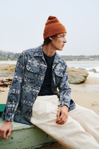 Man in patterned shirt and beanie poses seated outdoors on a beach with rocks, and ocean waves in the background on an overcast day.