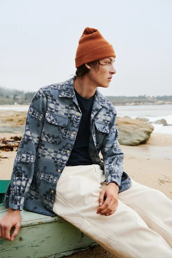 Man in patterned shirt and beanie poses seated outdoors on a beach with rocks, and ocean waves in the background on an overcast day.