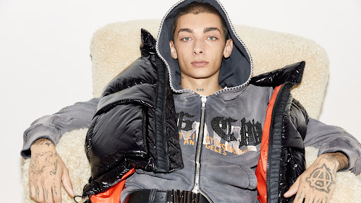 Portrait of a young adult male seated in a fuzzy chair, wearing a hooded sweatshirt, a black puffer vest, and a black belt. Tattoos are visible.