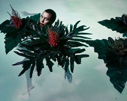 Artistic image of a woman immersed in water, surrounded by tropical foliage and red flowering plants in a surreal, dreamy setting.