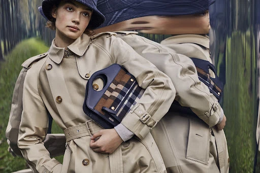 Adult female fashion model in a beige trench coat and hat, holding a handbag in an outdoor setting with a blurred background.
