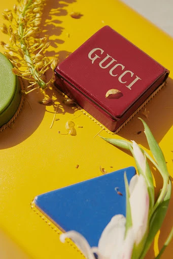 Close-up of a red Gucci box surrounded by a yellow surface, small flowers, a green container, and a blue triangle.