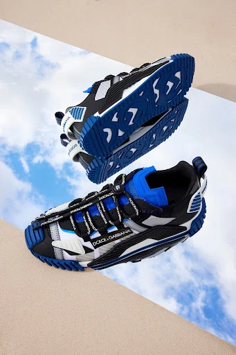 Two Dolce & Gabbana sneakers are displayed on a tan surface with a reflective surface, against a backdrop of blue skies and clouds.
