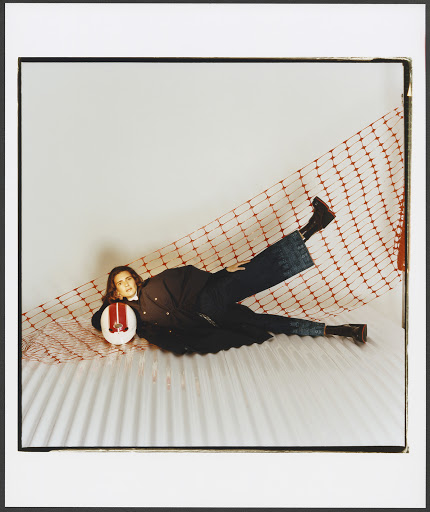 An adult female lies in a red-and-white net, wearing a black jacket and jeans, resting her head on a white helmet in a photo studio.