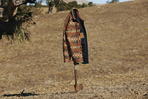 A patterned shirt-jacket hangs on a shovel in a rural field with dry grass and distant mountains on a sunny day.