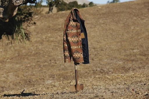 A patterned shirt-jacket hangs on a shovel in a rural field with dry grass and distant mountains on a sunny day.