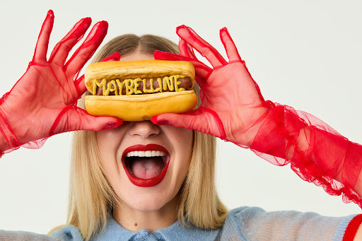 Blonde woman with wide smile in red gloves holds hotdog with 'Maybelline' written in mustard over her eyes in a bright studio.